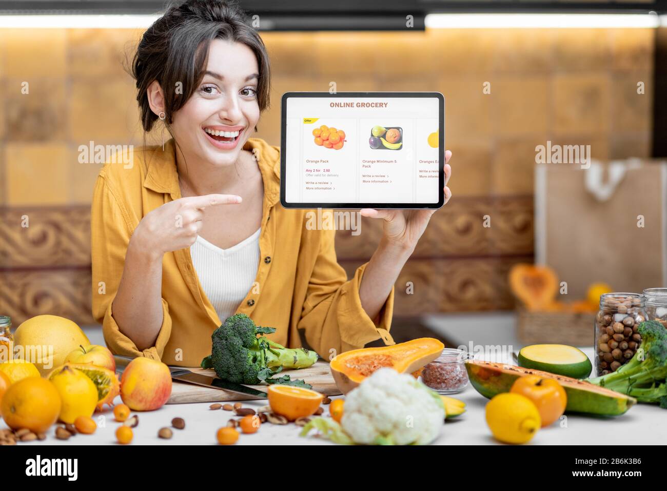 Femme tenant une tablette numérique avec le marché de shopping en ligne lancé tout en se tenant sur la cuisine avec beaucoup de nourriture fraîche sur la table Banque D'Images