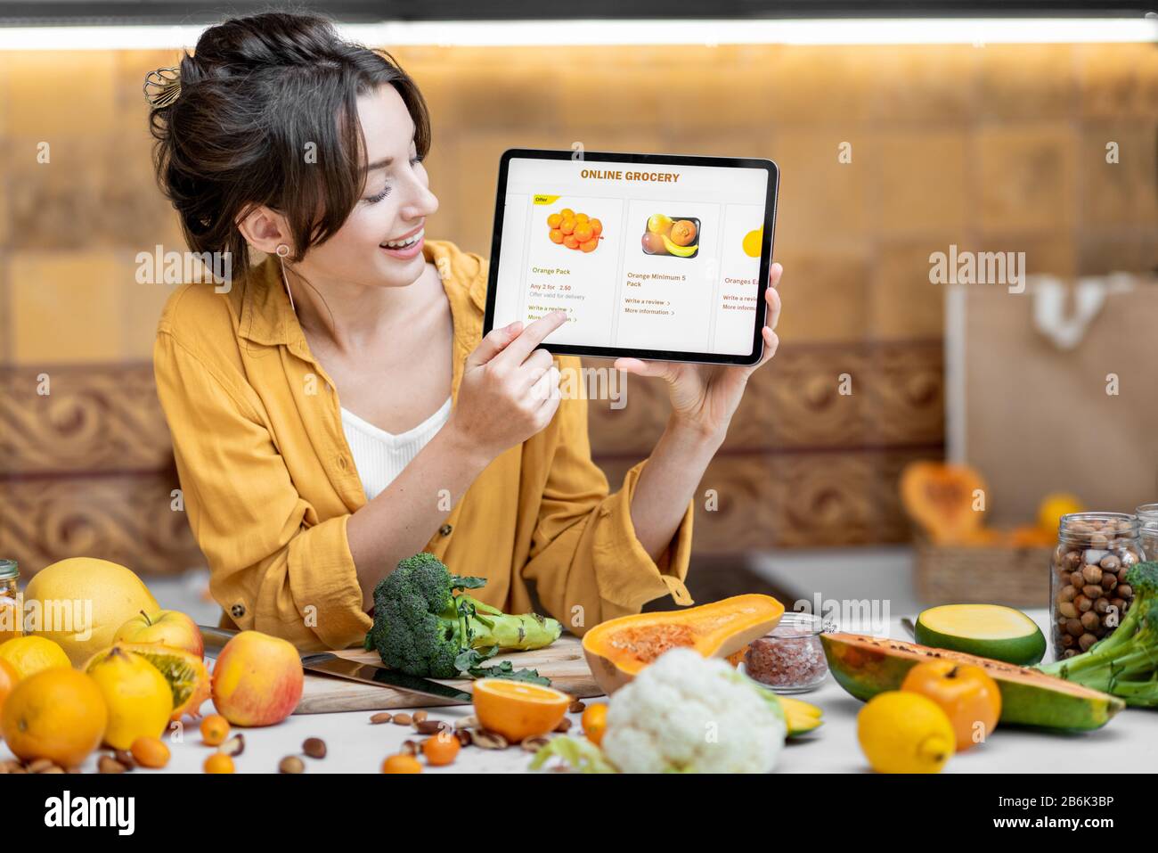 Femme tenant une tablette numérique avec le marché de shopping en ligne lancé tout en se tenant sur la cuisine avec beaucoup de nourriture fraîche sur la table Banque D'Images