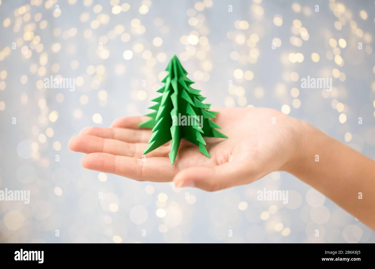 vacances d'hiver, nouvelle année et concept d'artisanat - main tenant le papier vert origami arbre de noël sur fond de lumières de fête Banque D'Images
