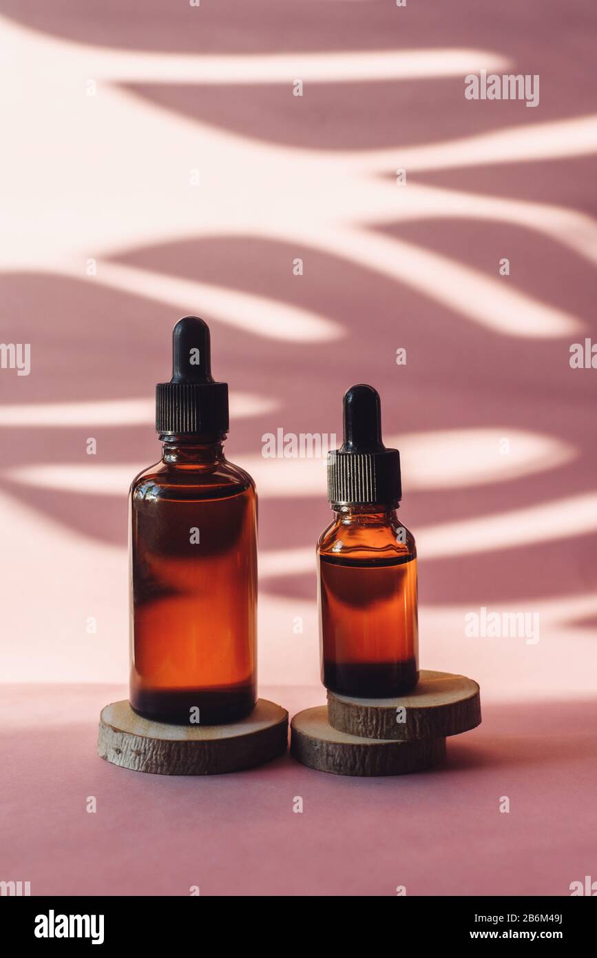 Deux bouteilles de verre avec sérum hydratant sur des découpes de scie en bois sur un fond rose. Tubes bruns avec compte-gouttes sur le fond d'une ombre tropicale d'une feuille de palmier. Le concept de soins de la peau. Banque D'Images