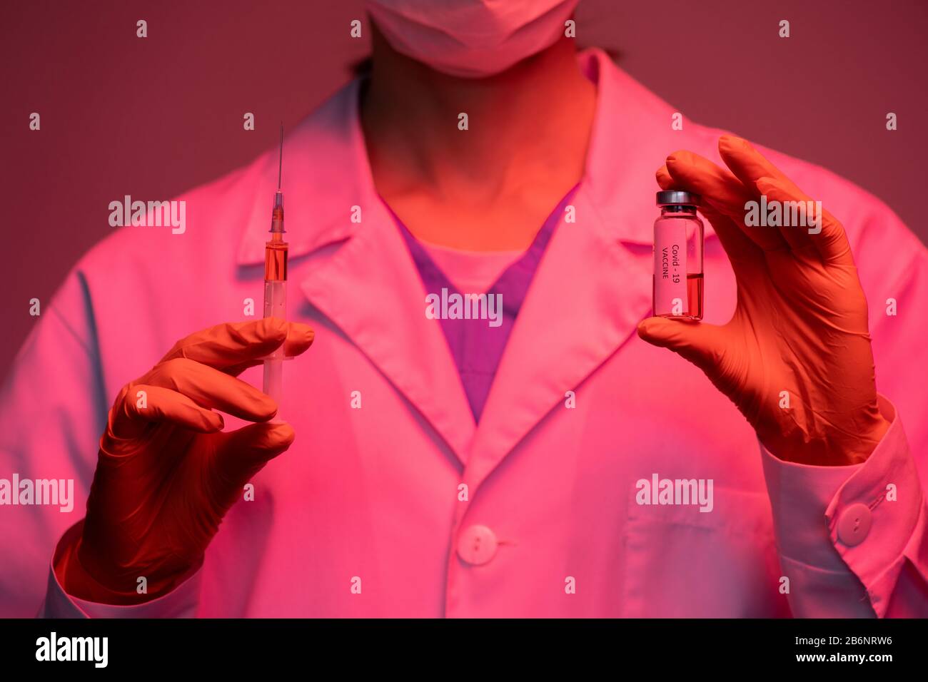 Gros plan de spécialiste médical non reconnaissable en gants contenant une bouteille de vaccin et une seringue Covid-19 en lumière rouge Banque D'Images