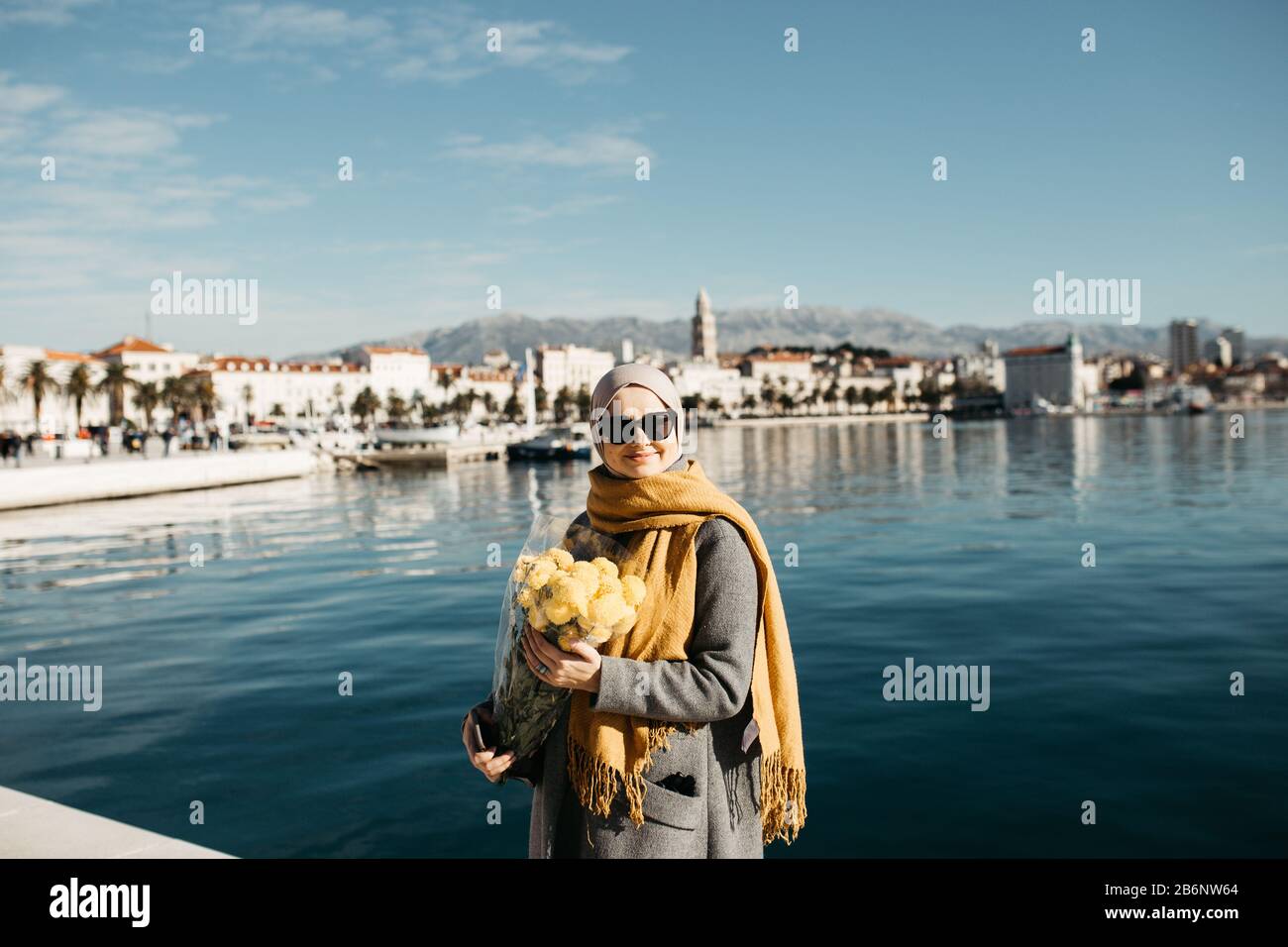 Portrait des femmes musulmanes européennes avec hijab assis sur la plage de pierre avec mer et port en arrière-plan. Elle tient le bouquet de fleurs jaunes. Banque D'Images