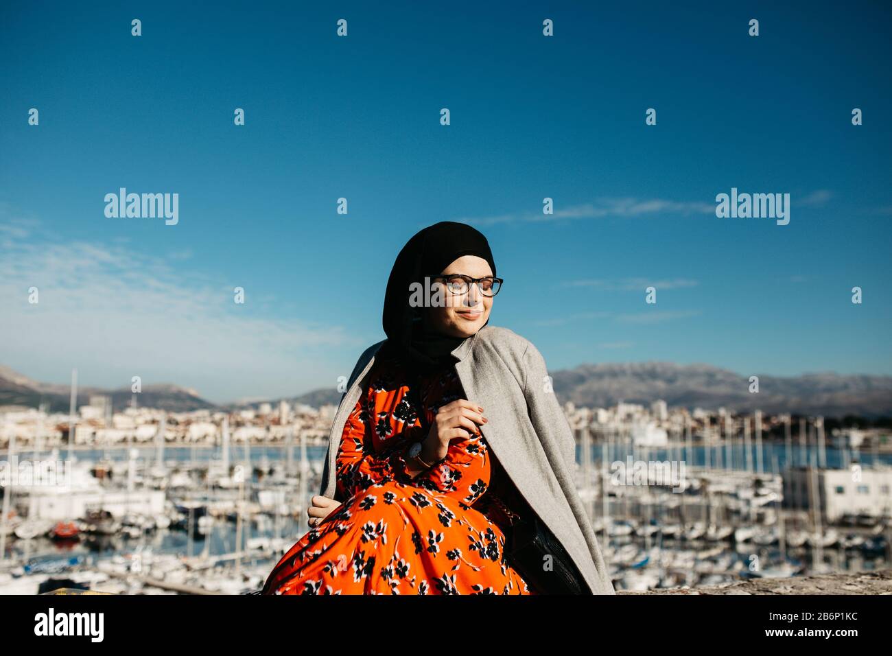 Portrait de jeunes femmes musulmanes européennes avec hijab assis sur la plage de pierre avec mer et port en arrière-plan. Elle est heureuse et profite du soleil. Banque D'Images