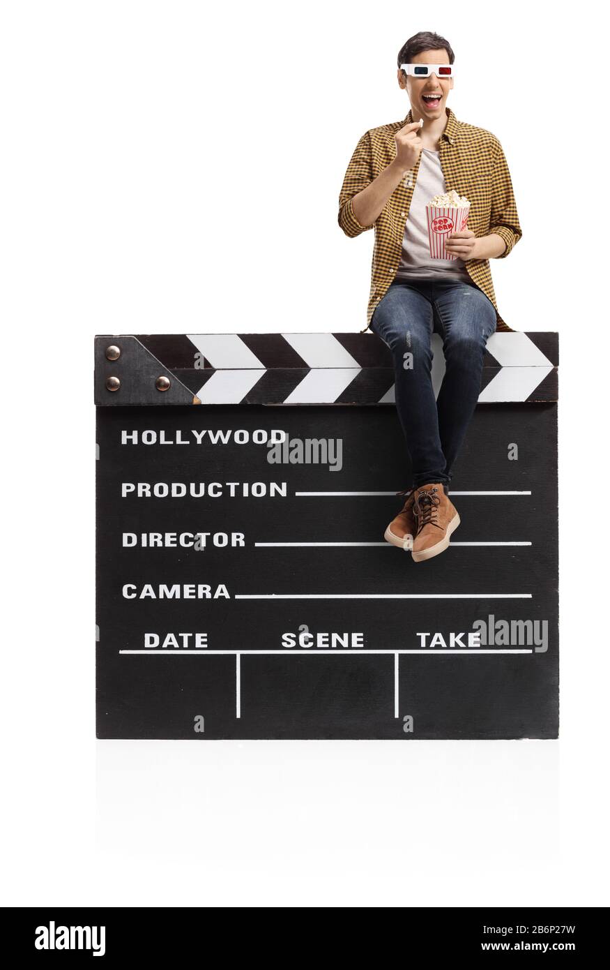 Jeune homme avec des lunettes en relief assis sur un panneau de clapper de film et manger du popcorn isolé sur fond blanc Banque D'Images