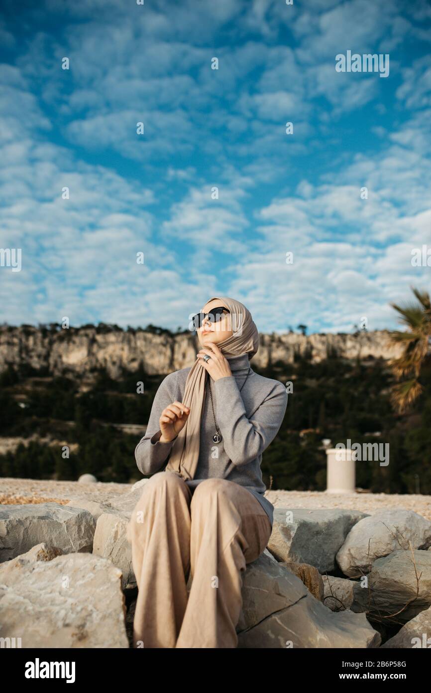 Portrait de jeunes femmes musulmanes européennes avec hijab assis sur la plage en pierre avec la mer en arrière-plan. Il est heureux et détendu. Banque D'Images