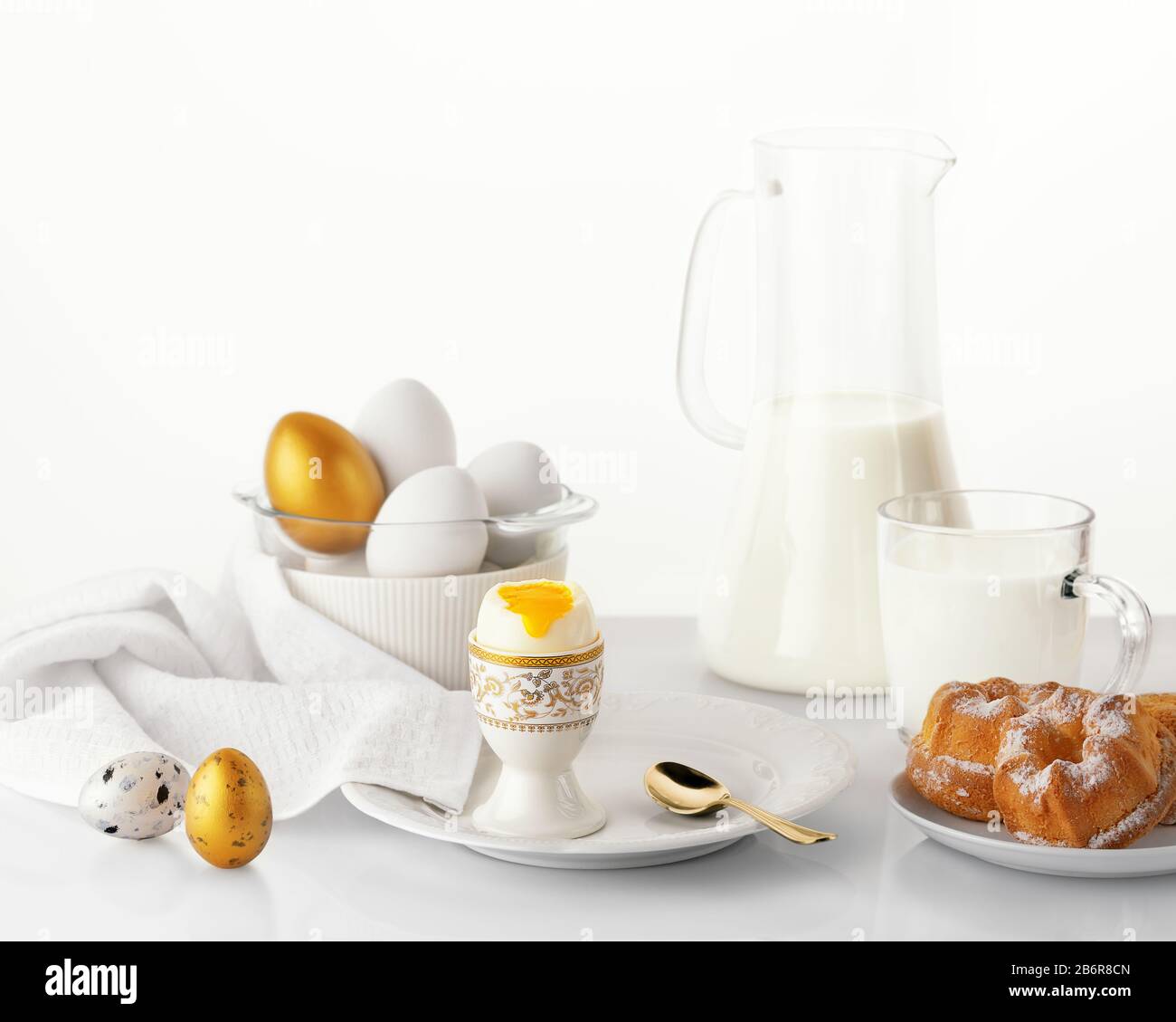 Oeuf cuit doux sur une plaque blanche avec des œufs de Pâques or et blanc, des œufs de caille, de la boulangerie et du lait sur fond blanc. Concept de petit déjeuner de Pâques. Banque D'Images