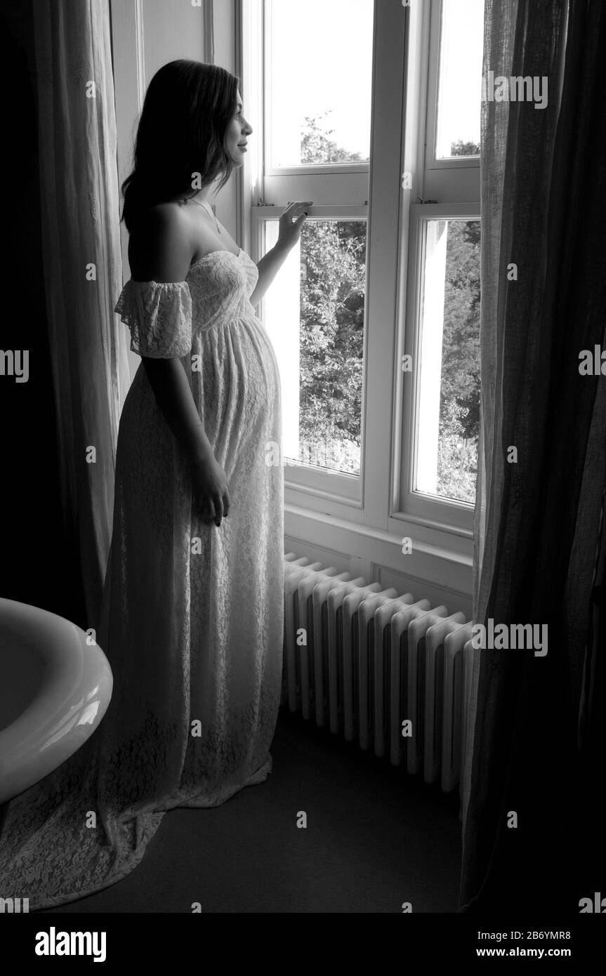 Belle jeune femme enceinte debout par fenêtre regardant sereine Banque D'Images