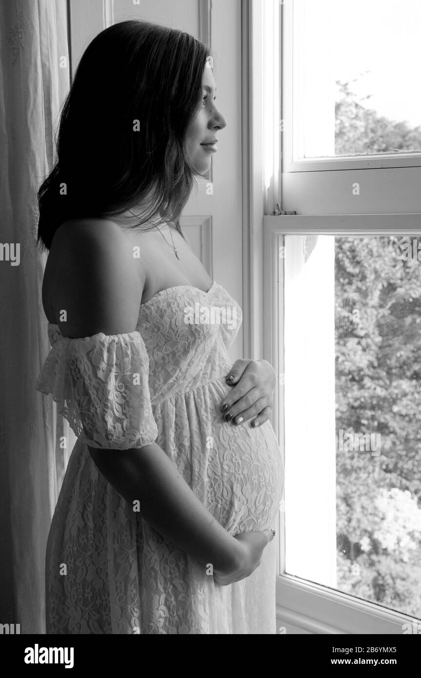 Belle jeune femme enceinte debout par fenêtre regardant sereine Banque D'Images