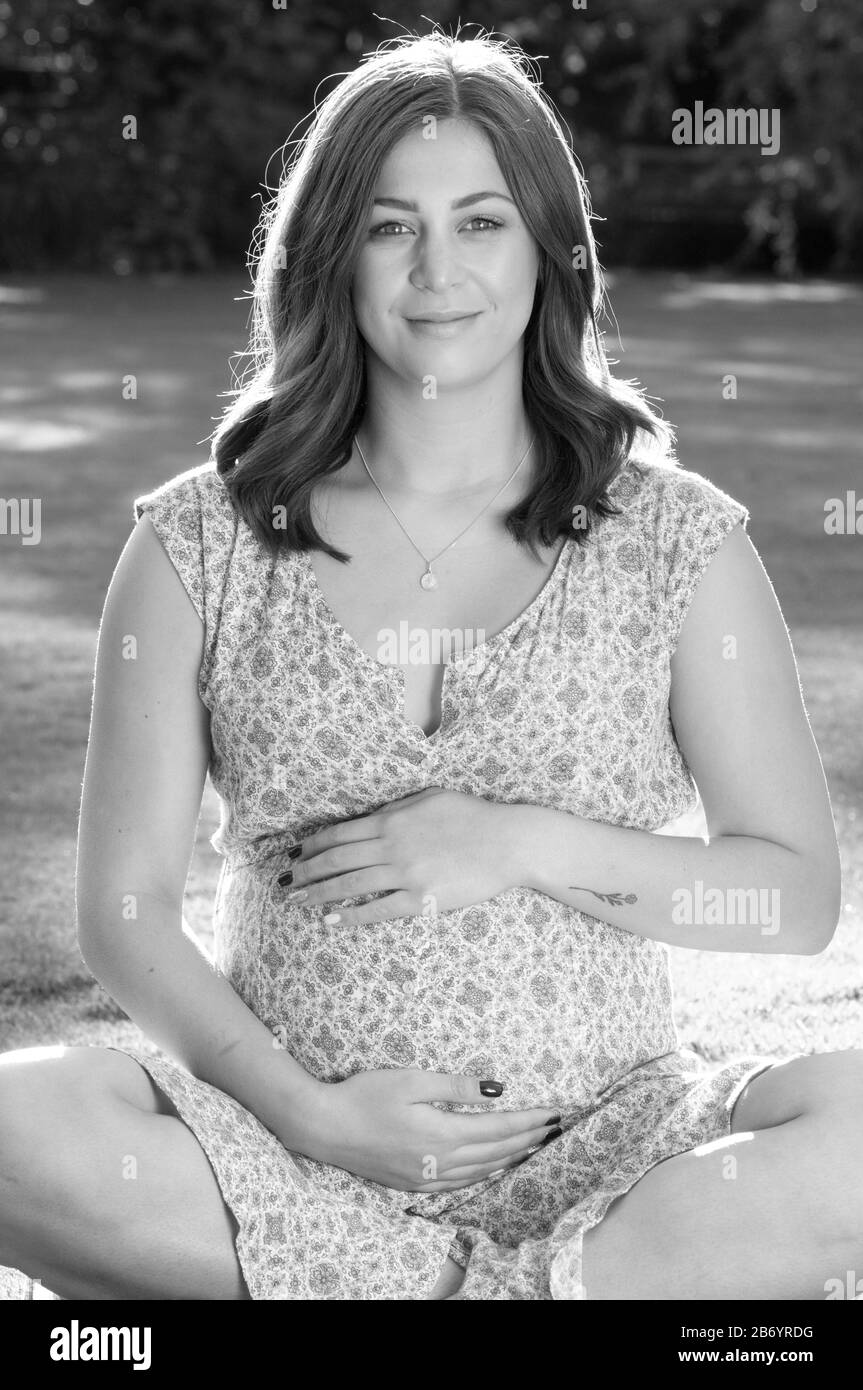 Portrait noir et blanc de la belle jeune femme enceinte assise sur l'herbe caressant son bosse de bébé Banque D'Images