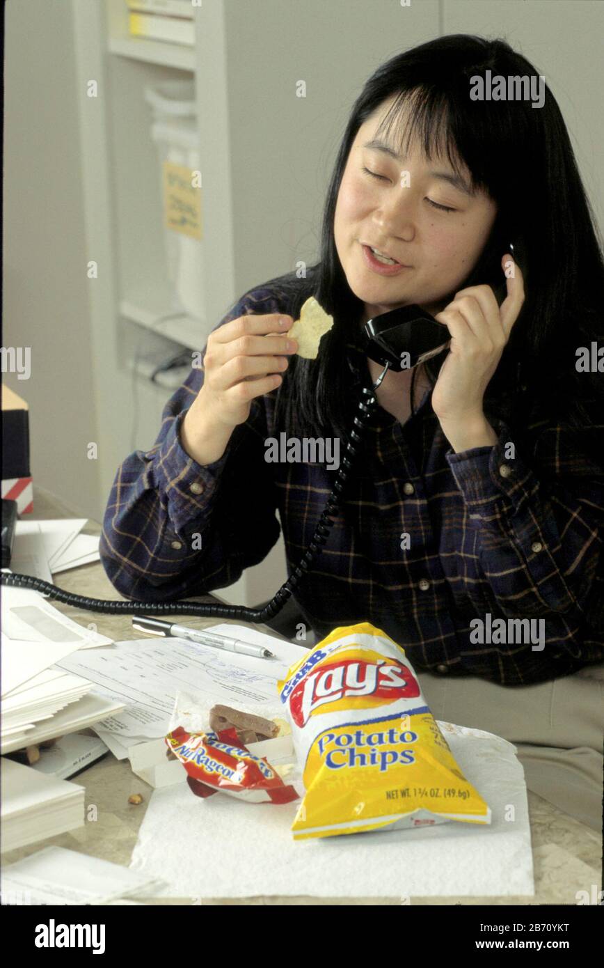 Habitudes alimentaires malsaines. La jeune femme américano-asiatique saute le déjeuner au travail et mange des chips de collation à la place. M. ©Bob Daemmrich Banque D'Images