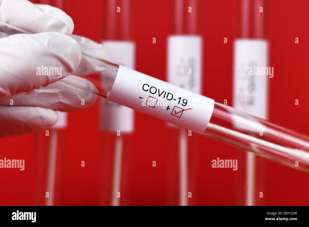Échantillon de tube d'essai médical positif pour le Coronavirus avec l'étiquette Covid-19 tenue à la main dans le gant en caoutchouc avec d'autres tubes de furie en fond rouge Banque D'Images