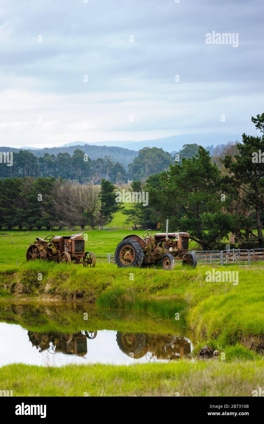 Yarra Valley, Victoria avec des prairies vallonnées, des terres boisées et deux tracteurs de ferme garés sur un mur de barrage avec leur réflexion dans l'eau. Banque D'Images