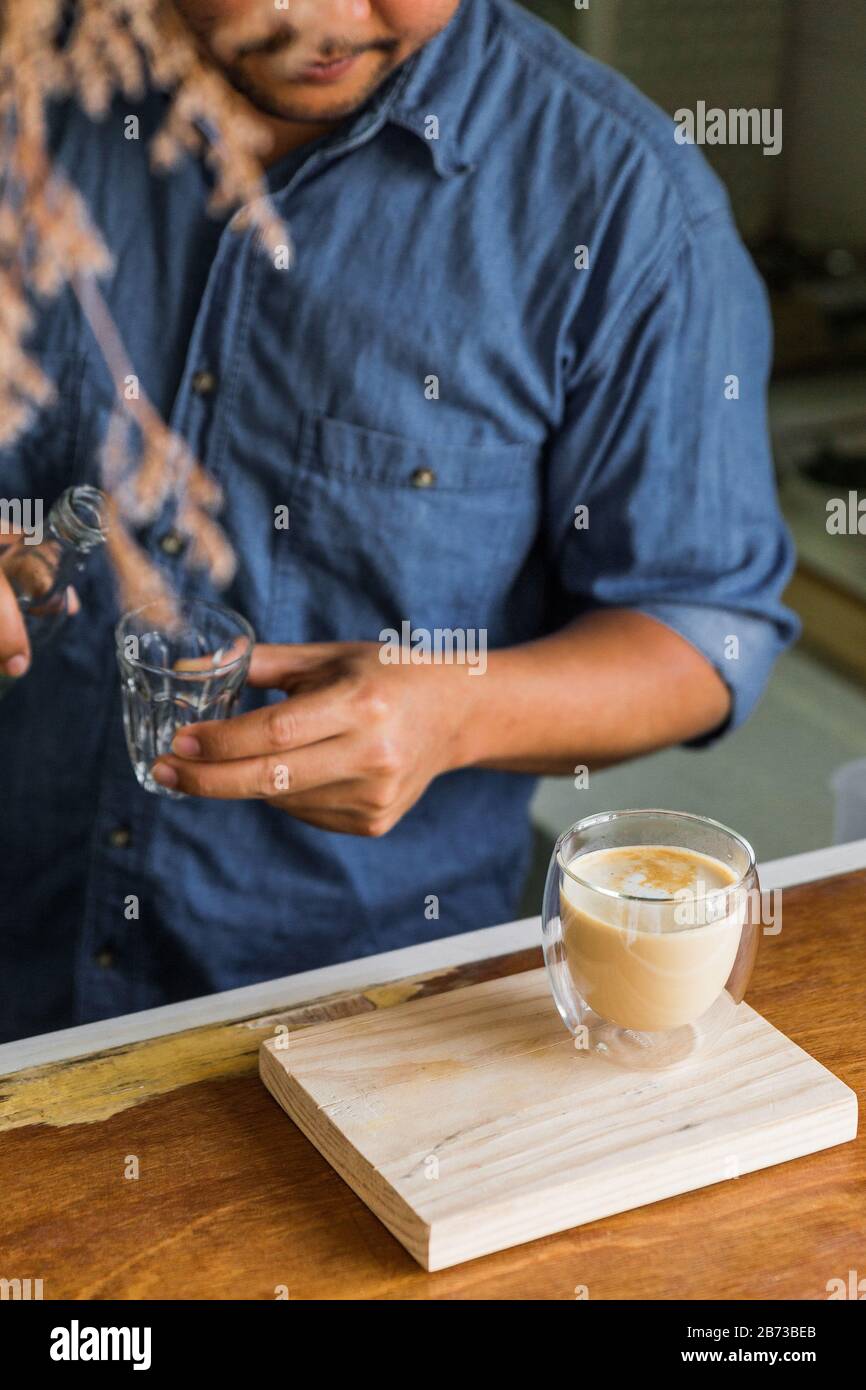 Barista masculin prépare de la mousse de lait dans du café au Latte chaud dans du verre à boire au comptoir en bois. Servi sur une plaque en bois. Banque D'Images