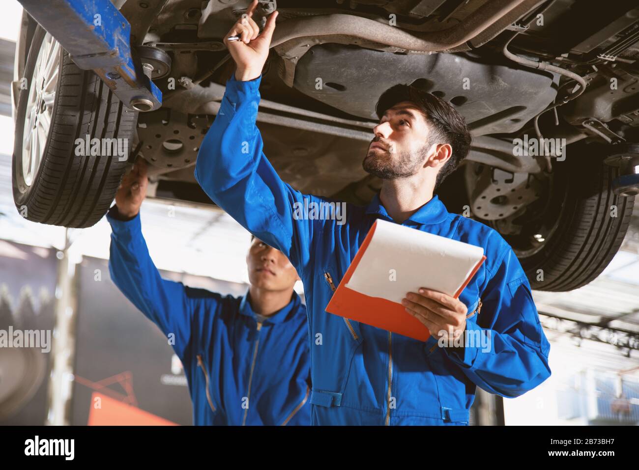 Le mécanicien en uniforme bleu d'usure inspecte le fond de la voiture avec son assistant. Service de réparation automobile, travail d'équipe professionnel. Banque D'Images