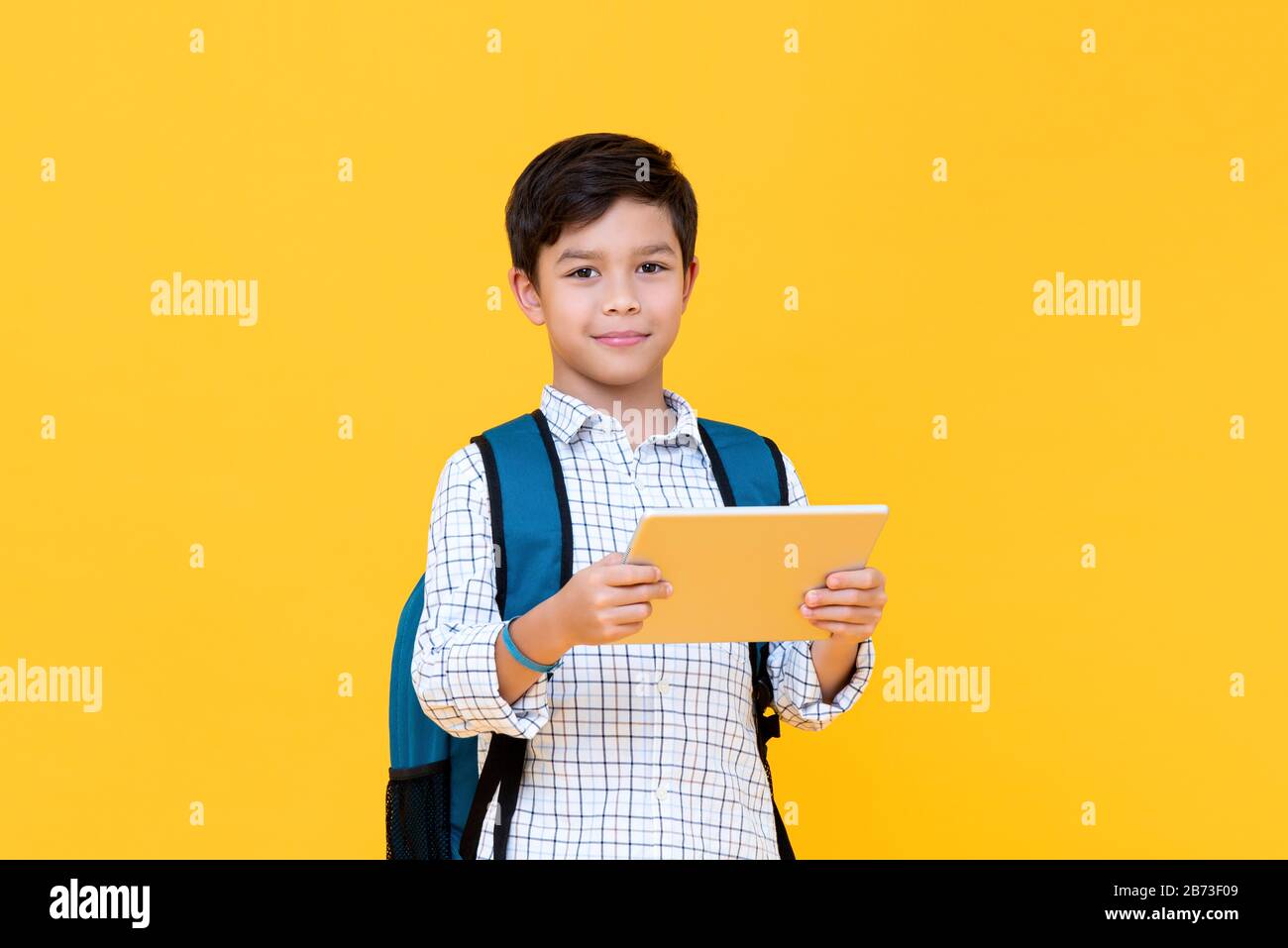 Taille haut portrait de beau jeune écolier portant sac à dos tenant une tablette tout en regardant directement dans l'appareil photo dans l'arrière-plan jaune isolé de studio Banque D'Images