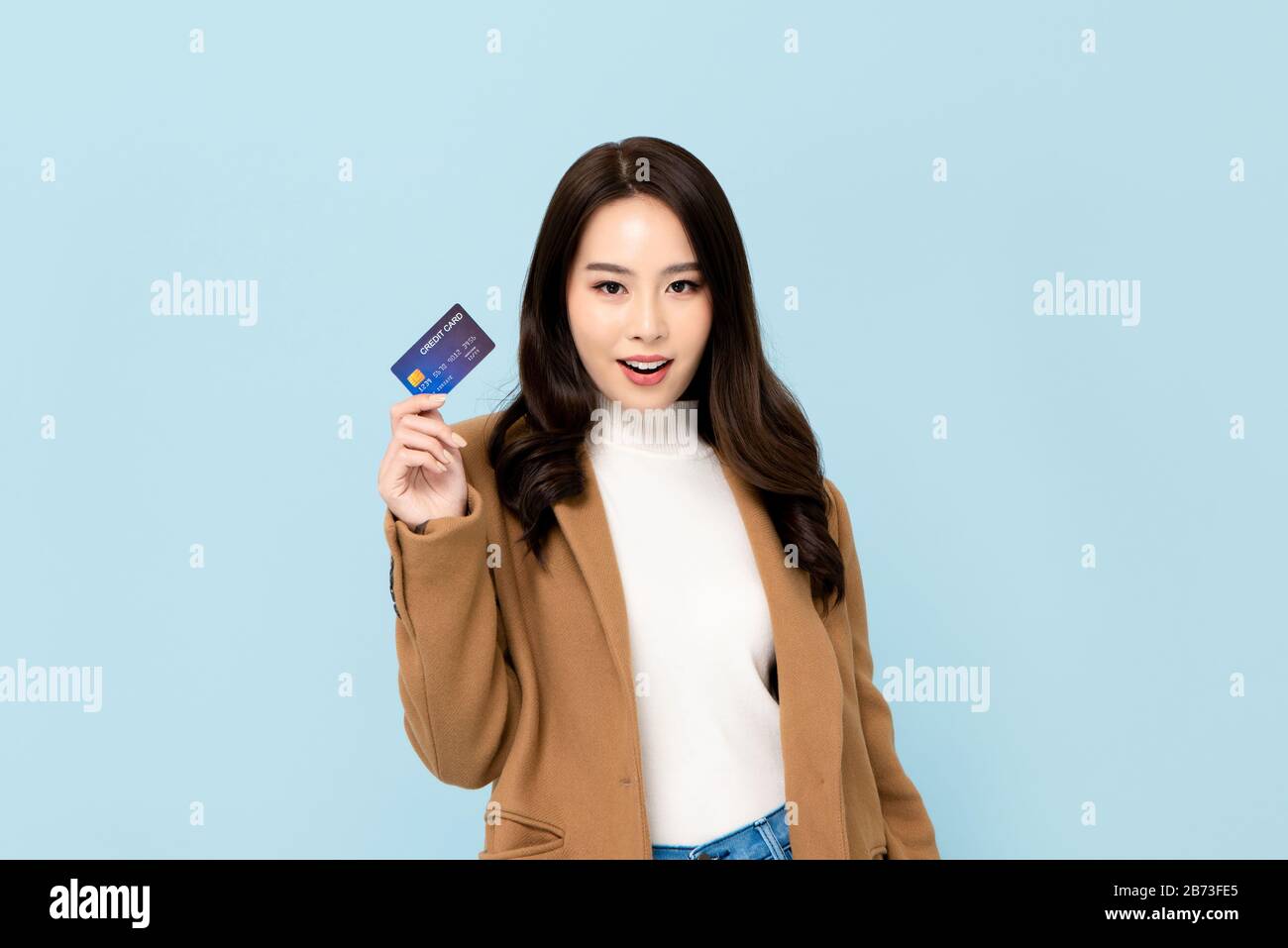 Belle femme asiatique souriante en vêtements d'hiver montrant carte de crédit en main pour les concepts financiers et de société sans cash isolés sur fond bleu clair Banque D'Images