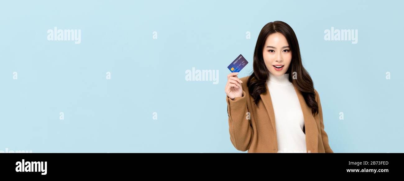 Belle femme asiatique souriante en vêtements d'hiver montrant carte de crédit en main pour les concepts financiers et de société sans cash isolés sur la bannière bleu clair Banque D'Images