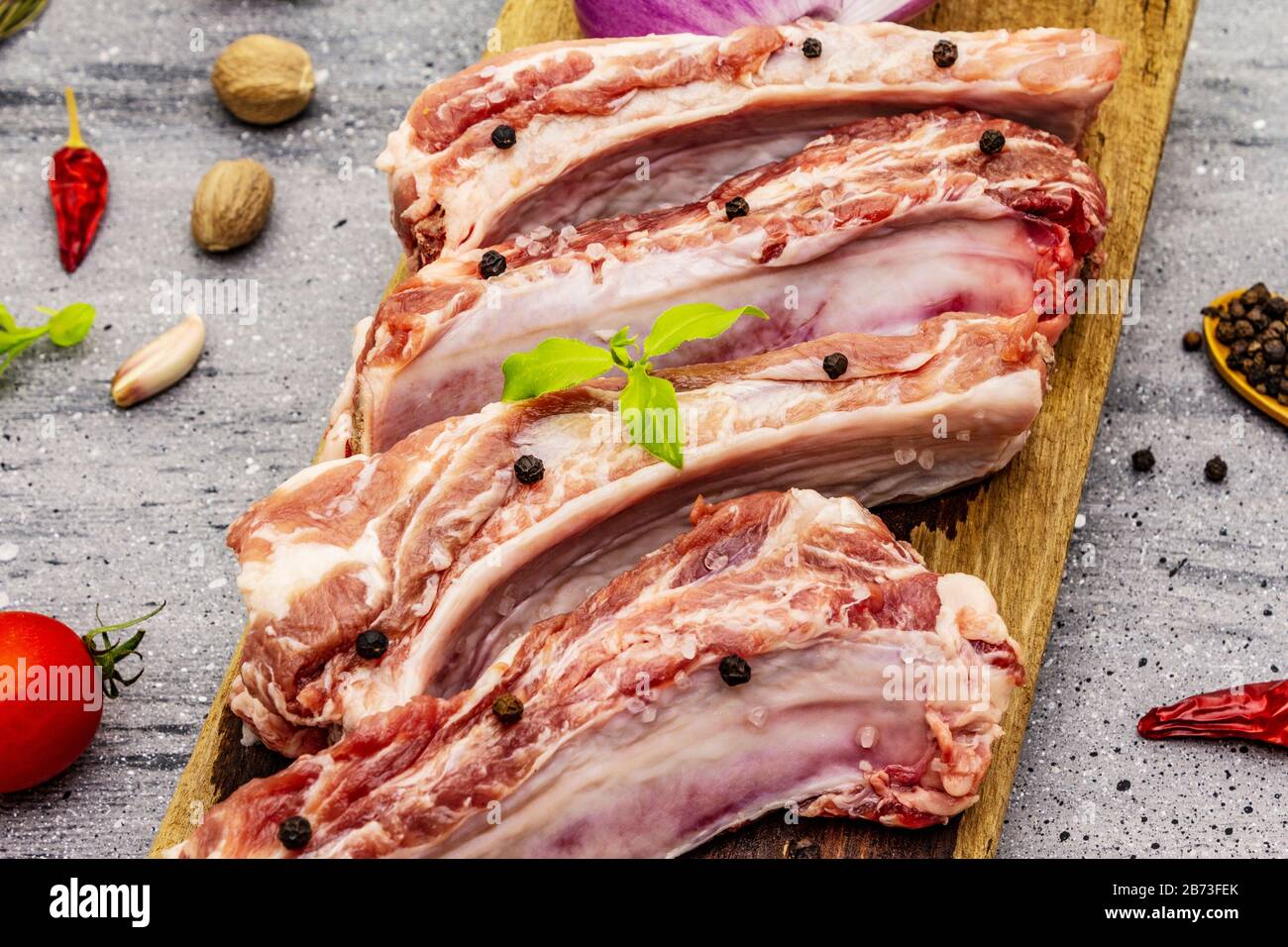 Côtes de porc brutes sur fond de béton gris. Planche à découper ronde en bois, légumes, épices et légumes verts Banque D'Images