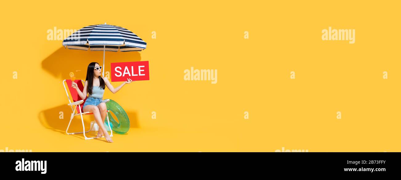 Surprise belle femme asiatique en été tenue tenant le signe de vente rouge tout en étant assis sur chaise de plage isolée sur fond de bannière jaune avec copie spa Banque D'Images