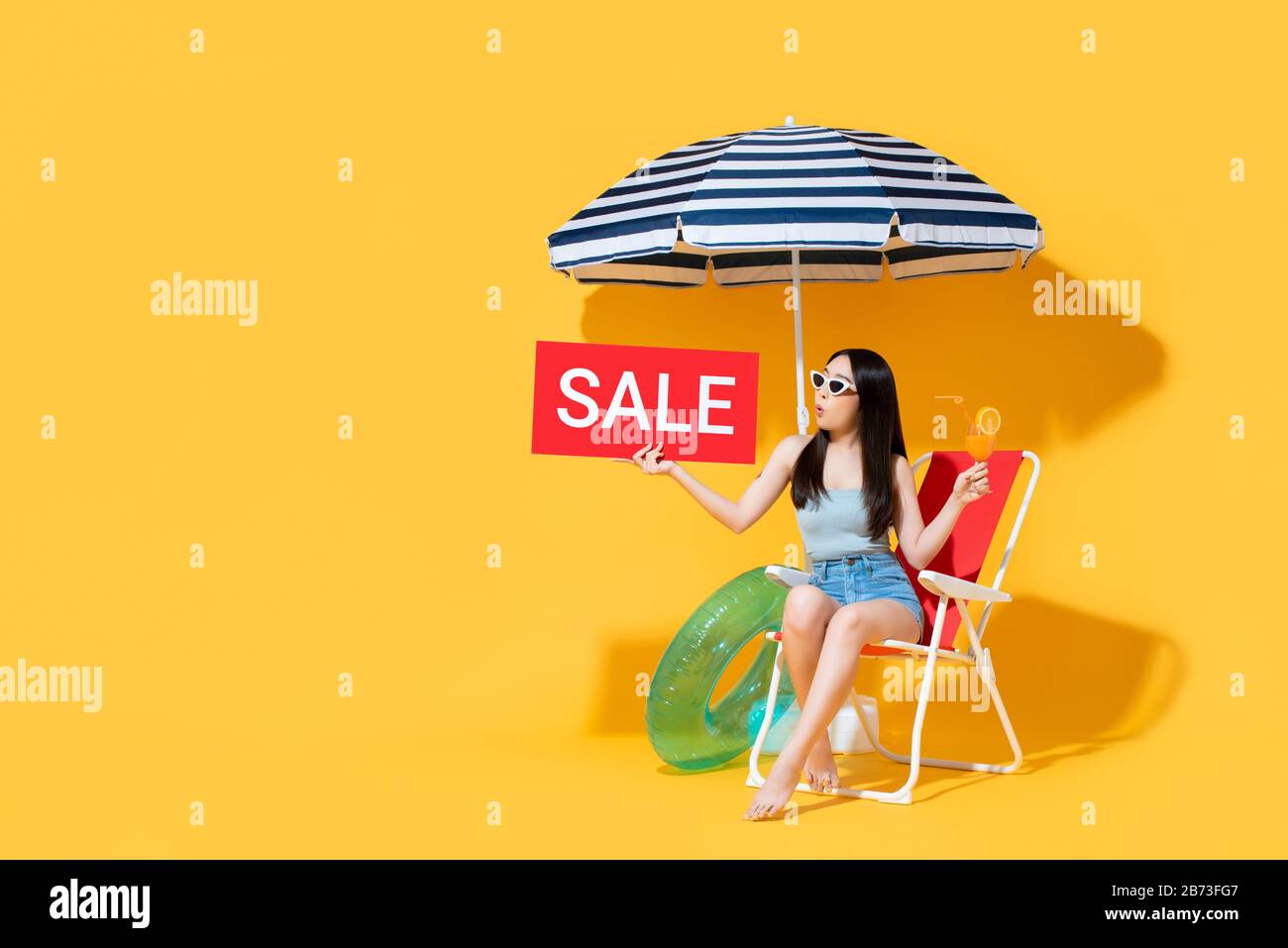 Surprise belle femme asiatique en été tenue tenant le signe de vente rouge tout en étant assis sur chaise de plage isolée sur fond jaune avec espace de copie Banque D'Images