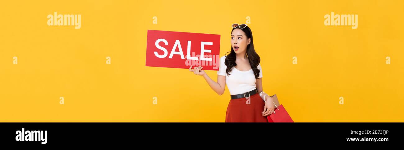 Choqué belle femme asiatique portant des sacs de shopping montrant le signe de vente rouge isolé sur fond de bannière jaune Banque D'Images