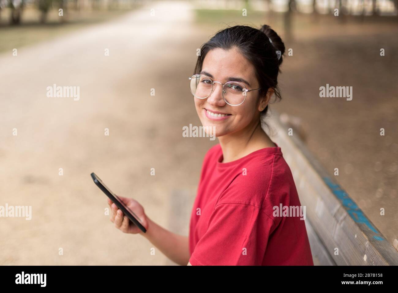 Jeune femme séduisante avec des verres assis sur un banc et utilisant son téléphone dans un parc, avec une chemise rouge. Séville, Espagne Banque D'Images