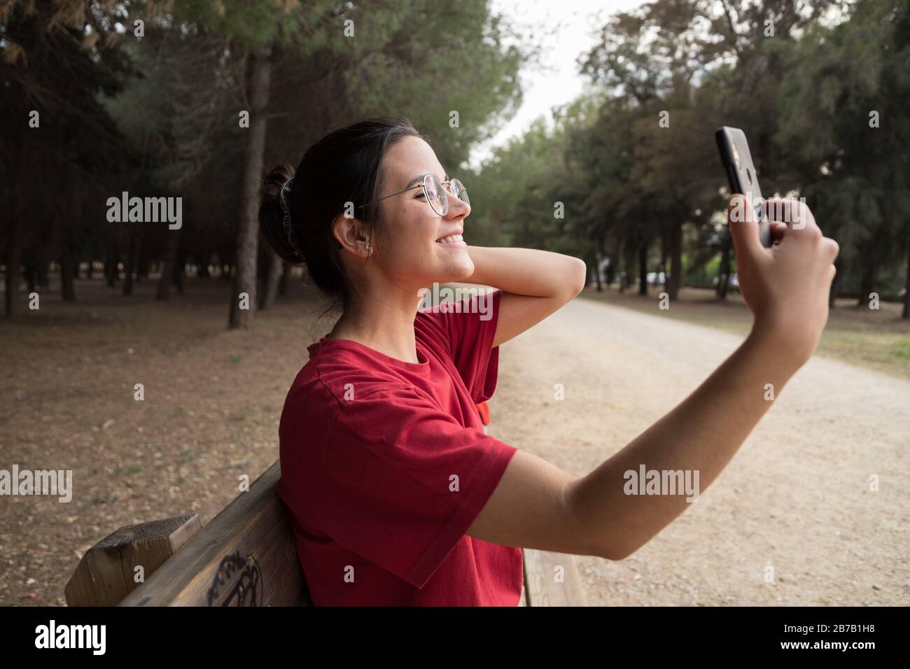 Jeune femme séduisante dans ses années vingt avec des verres et une chemise rouge assis sur un banc, tenant un téléphone mobile et prenant un selfie tout en souriant dans un Banque D'Images