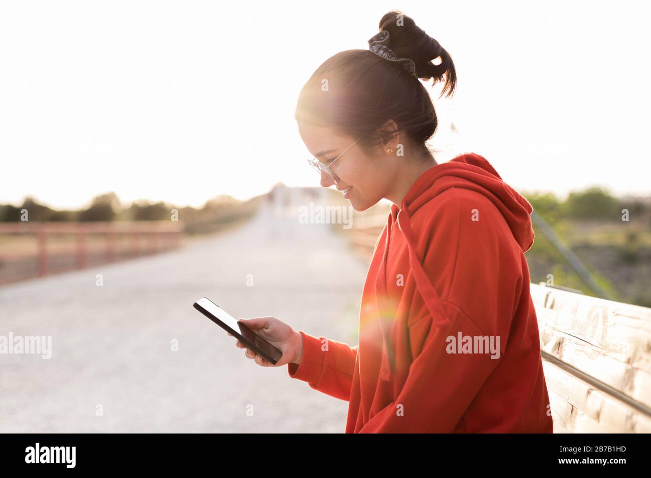 Jeune femme avec des lunettes sur ses années vingt discuter sur son téléphone tout en étant assis dans un banc d'un parc. Elle est souriante et porte un pull rouge et un poney Banque D'Images