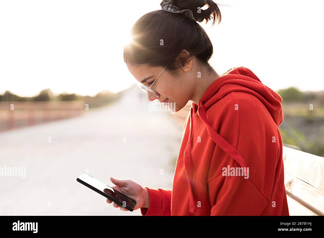Jeune femme avec des lunettes sur ses années vingt discuter sur son téléphone tout en étant assis dans un banc d'un parc. Elle est souriante et porte un pull rouge et un poney Banque D'Images
