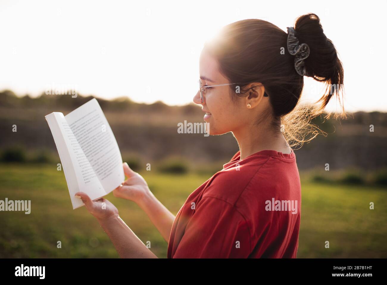 Jeune femme ayant une belle lecture tout en souriant et en profitant de la lumière d'un coucher de soleil dans un parc. Banque D'Images