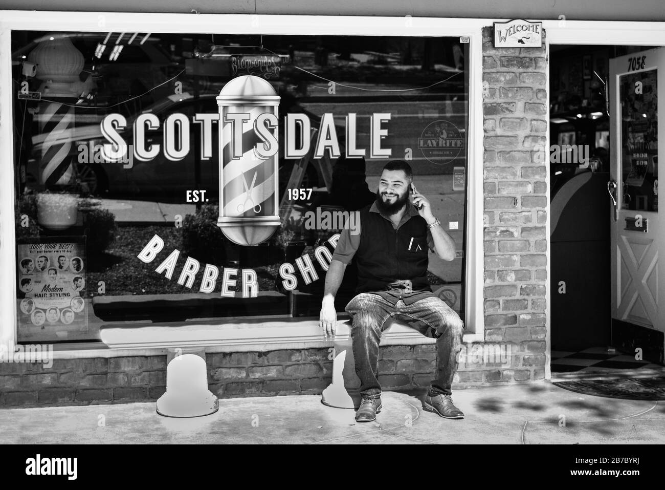 Un homme souriant, barbu, branché sur le téléphone portable est assis sur un banc devant le salon de coiffure Scottsdale, dans la vieille ville, Scottsdale, AZ, USA, en noir et blanc Banque D'Images