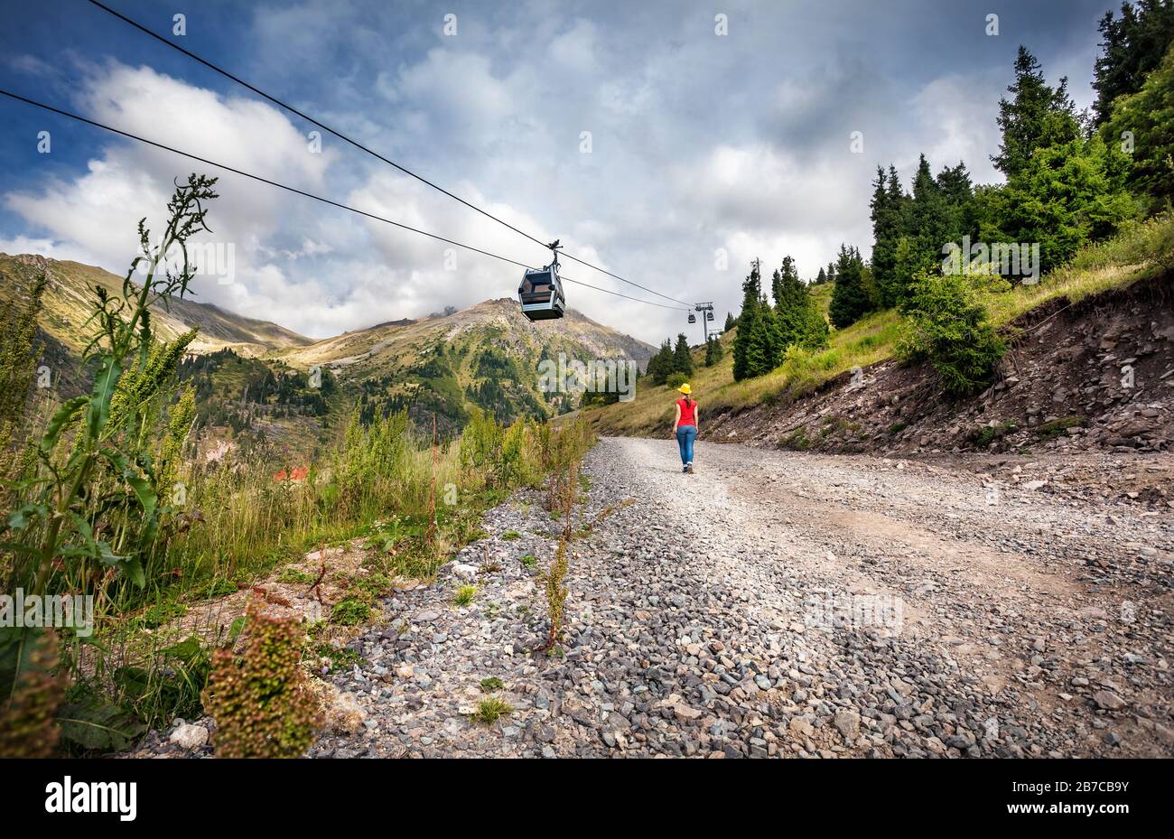 Femme de tourisme avec sac à dos et chapeau jaune marchant sur la route près du téléphérique dans la vallée de la montagne Banque D'Images