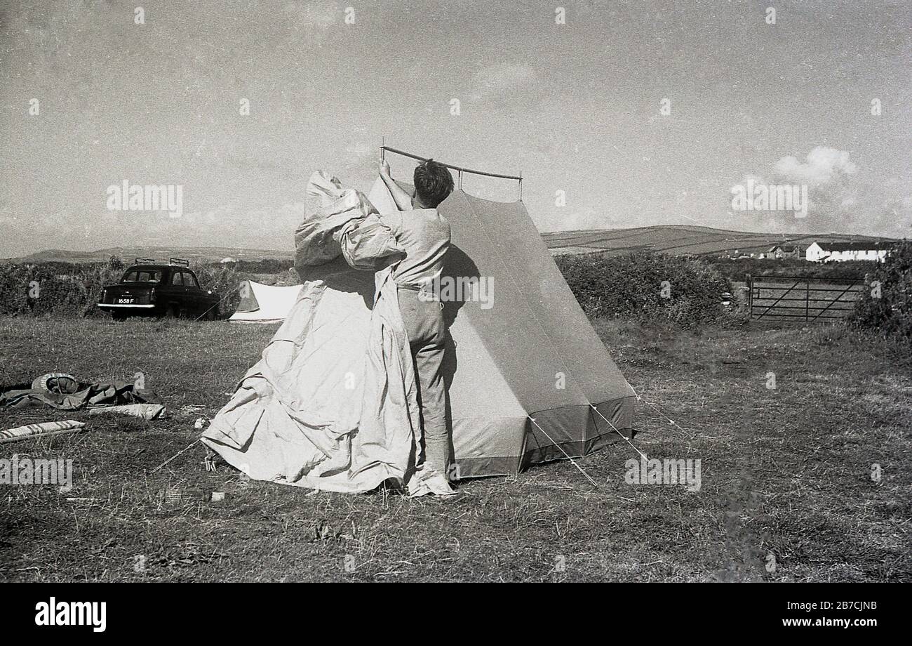 Camping à la côte style des années 1950, un campeur mâle dans un champ avec une couverture protectrice autour de lui mettant sa tente, Cornwall, Angleterre, Royaume-Uni. Une voiture de l'époque et une petite tente peuvent être vues à l'extrême bord du champ. Banque D'Images