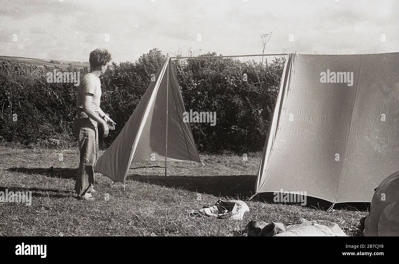 Camping à la côte style des années 1950, un campeur mâle dans un champ avec une couverture protectrice autour de lui mettant sa tente, Cornwall, Angleterre, Royaume-Uni. Une voiture de l'époque et une petite tente peuvent être vues à l'extrême bord du champ. Banque D'Images