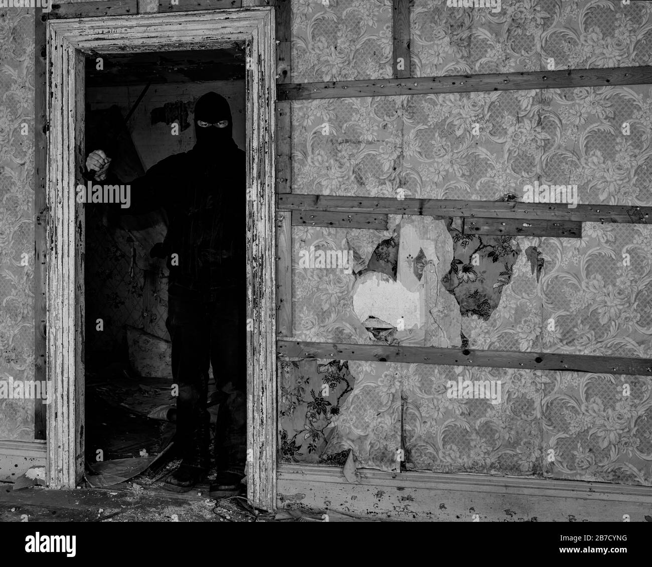 Un homme portant un masque/une capuche dans un bâtiment abandonné. Il tient un grand couteau, et se tient dans l'ombre dans une porte. Noir et blanc. Banque D'Images