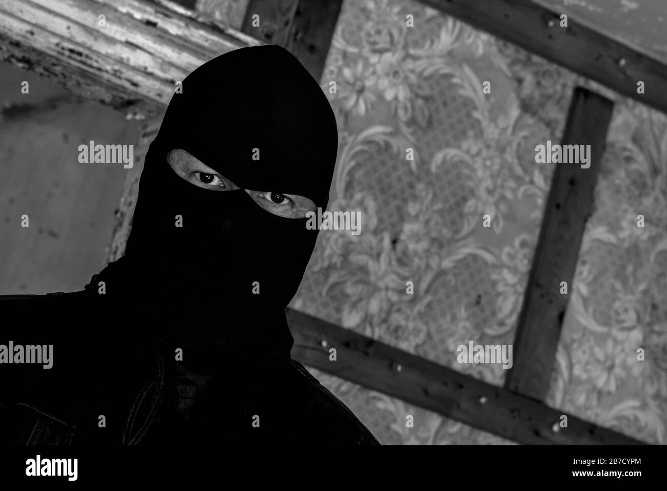 Un homme portant un masque/une capuche dans un bâtiment abandonné. Il est affamé au spectateur. Vue gros plan à un angle. Image noir et blanc foncé. Banque D'Images