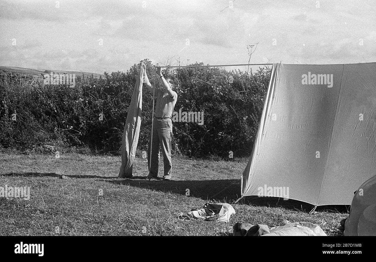Camping à la côte style des années 1950, un campeur mâle dans un champ avec une couverture protectrice élevée autour de lui mettant sa tente, Cornwall, Angleterre, Royaume-Uni. Banque D'Images
