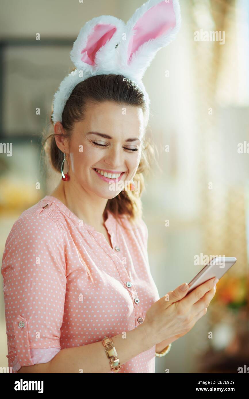 femme souriante d'âge moyen dans une blouse rose et des oreilles de lapin de pâques dans la maison moderne en journée ensoleillée de printemps écrire sms. Banque D'Images