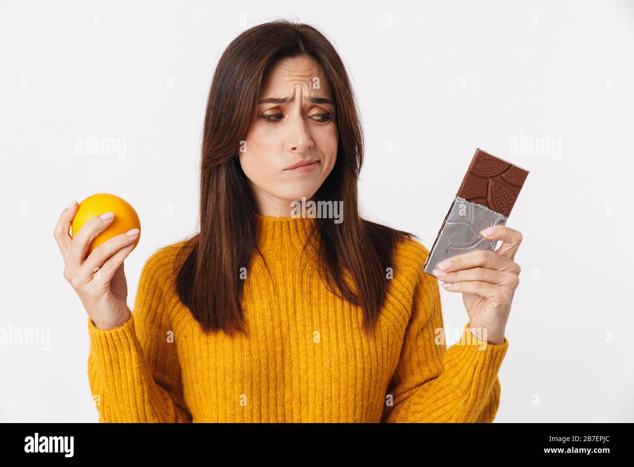 Image d'une femme adulte douteuse de brunette hésitant tout en tenant une barre d'orange et de chocolat isolée sur fond blanc Banque D'Images