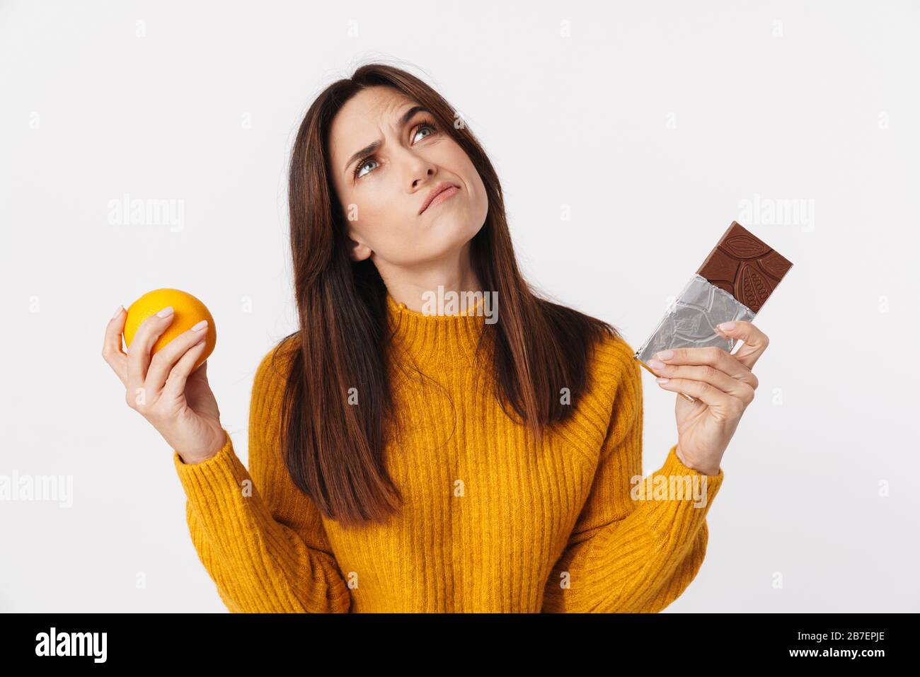 Image d'une femme adulte douteuse de brunette hésitant tout en tenant une barre d'orange et de chocolat isolée sur fond blanc Banque D'Images