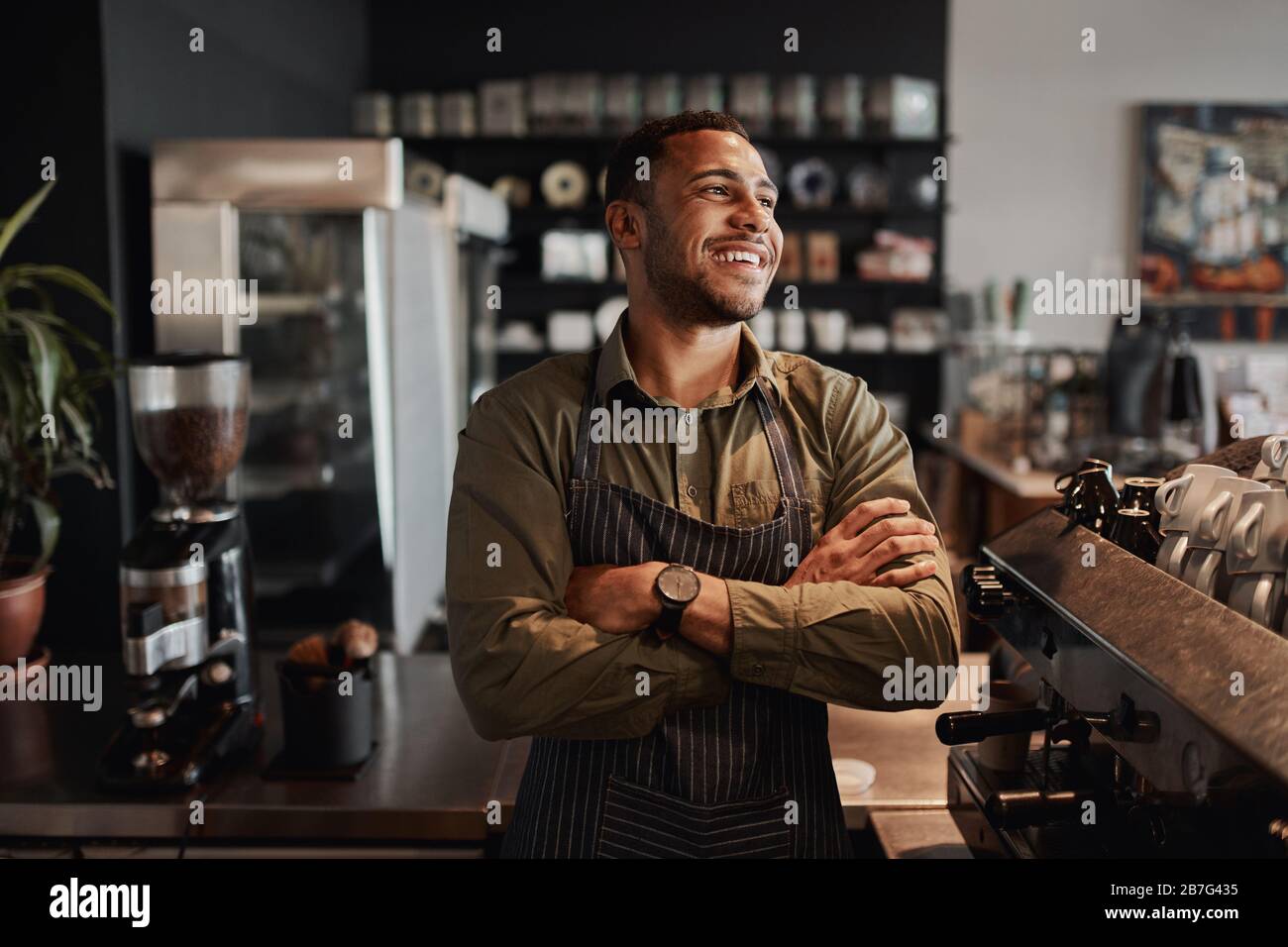 Jeune propriétaire afro-américain de petit café debout derrière le comptoir portant un tablier avec des bras croisés regardant loin Banque D'Images