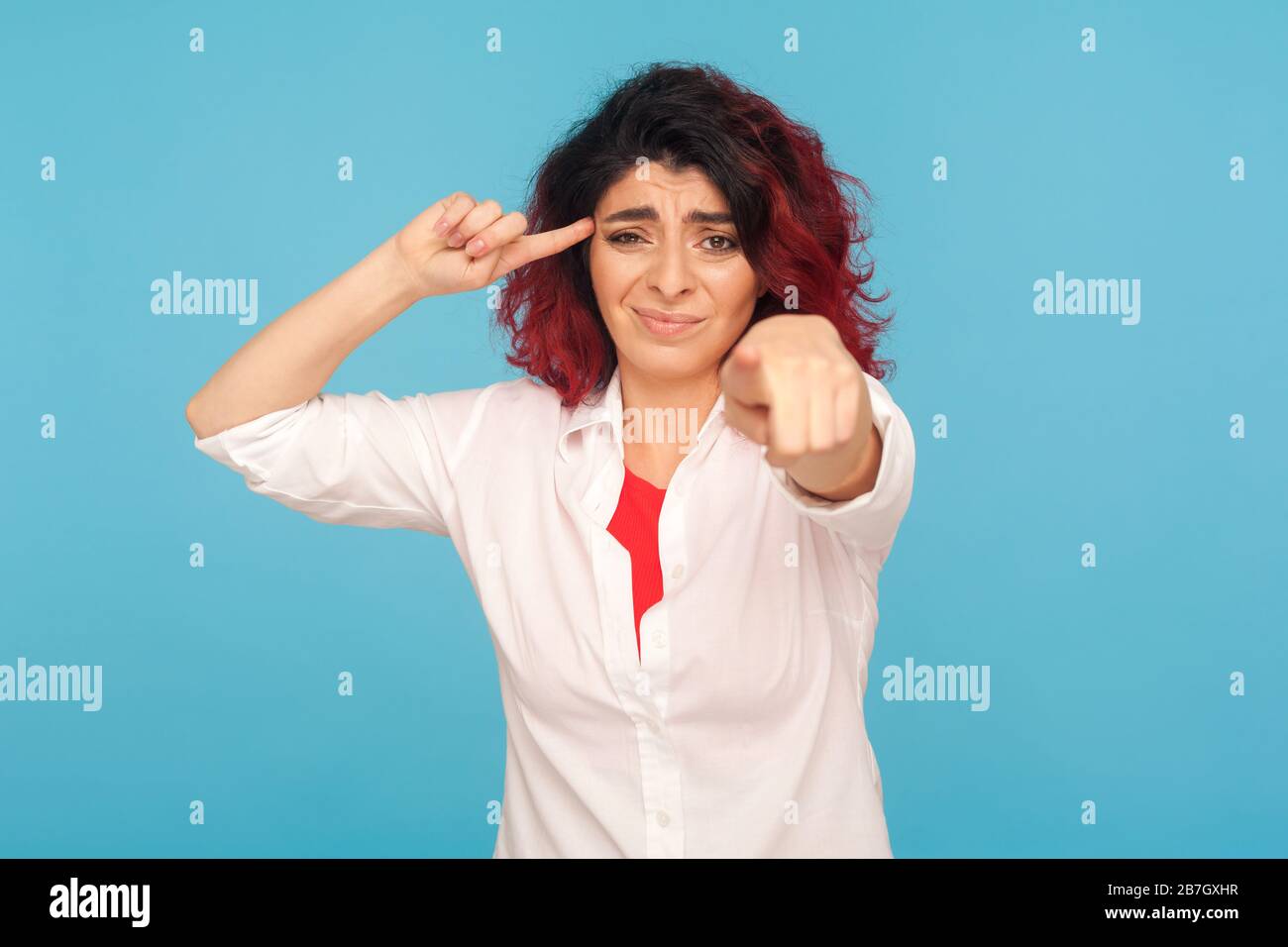 Votre idée stupide. Portrait d'une femme hipster avec des cheveux rouges fantaisie montrant un geste de couckoo stupide et pointant vers l'appareil photo, insatisfait de parler insensé. Banque D'Images