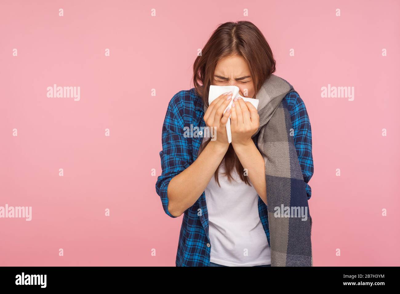 Grippe saisonnière. Portrait de la jeune fille malade dans une chemise à carreaux enveloppée dans un foulard soufflant le nez liquide, éternuant dans la serviette, souffrant de symptômes d'infection virale. In Banque D'Images