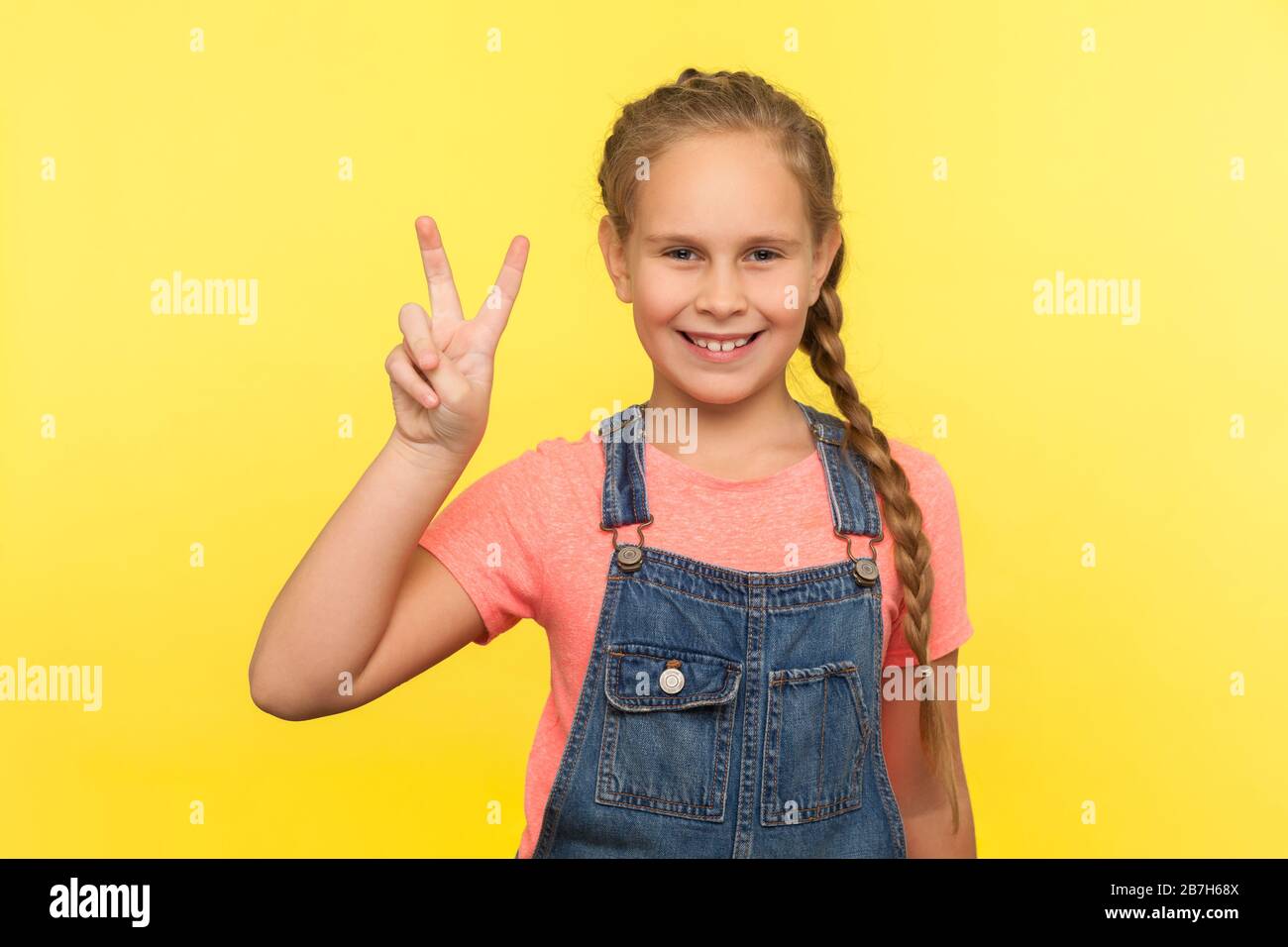 Portrait d'adorable petite fille positive avec tresse dans des salopettes en denim montrant le signe v avec les doigts doubles, la victoire gestuelle, célébrant le succès. ind Banque D'Images