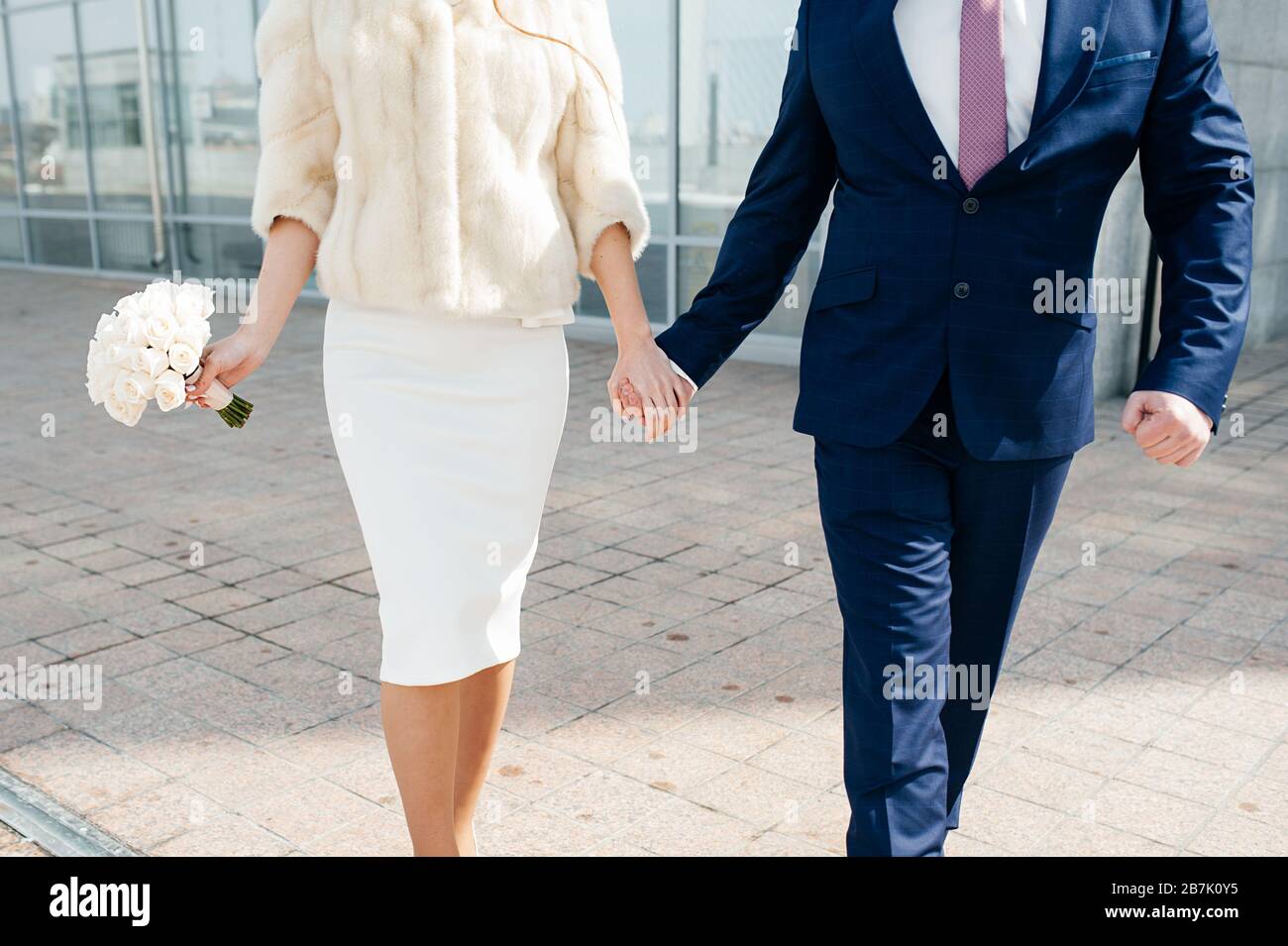 Le marié dans un costume bleu et la mariée dans une élégante robe de longueur de genou et un manteau de fourrure beige et un bouquet blanc de la mariée tiennent les mains et marchent sur le Banque D'Images