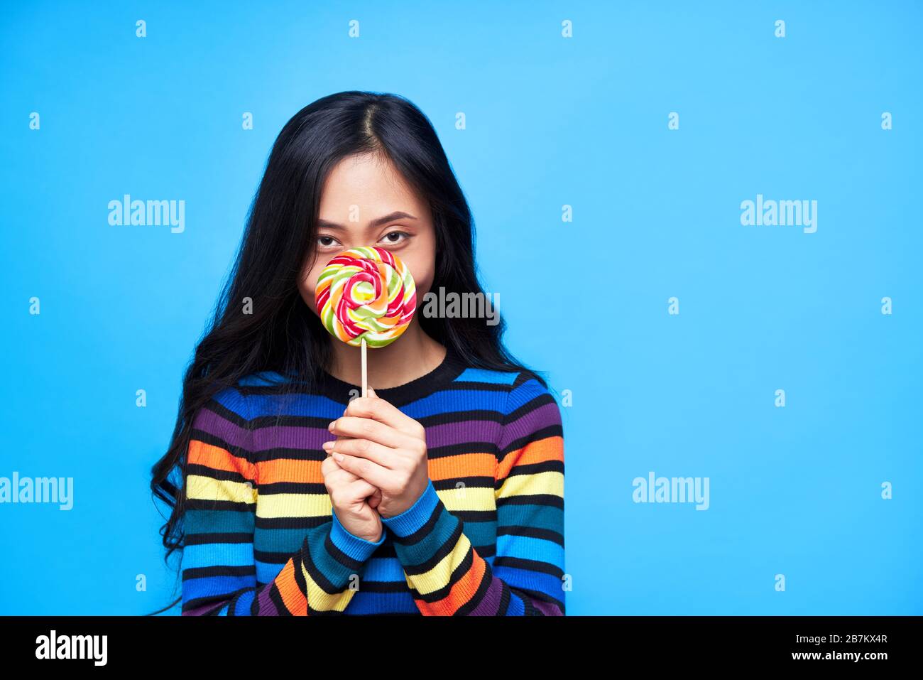 La jeune femme cache son visage derrière un lollipop coloré isolé sur fond bleu. Cocept d'émotion, nourriture douce Banque D'Images