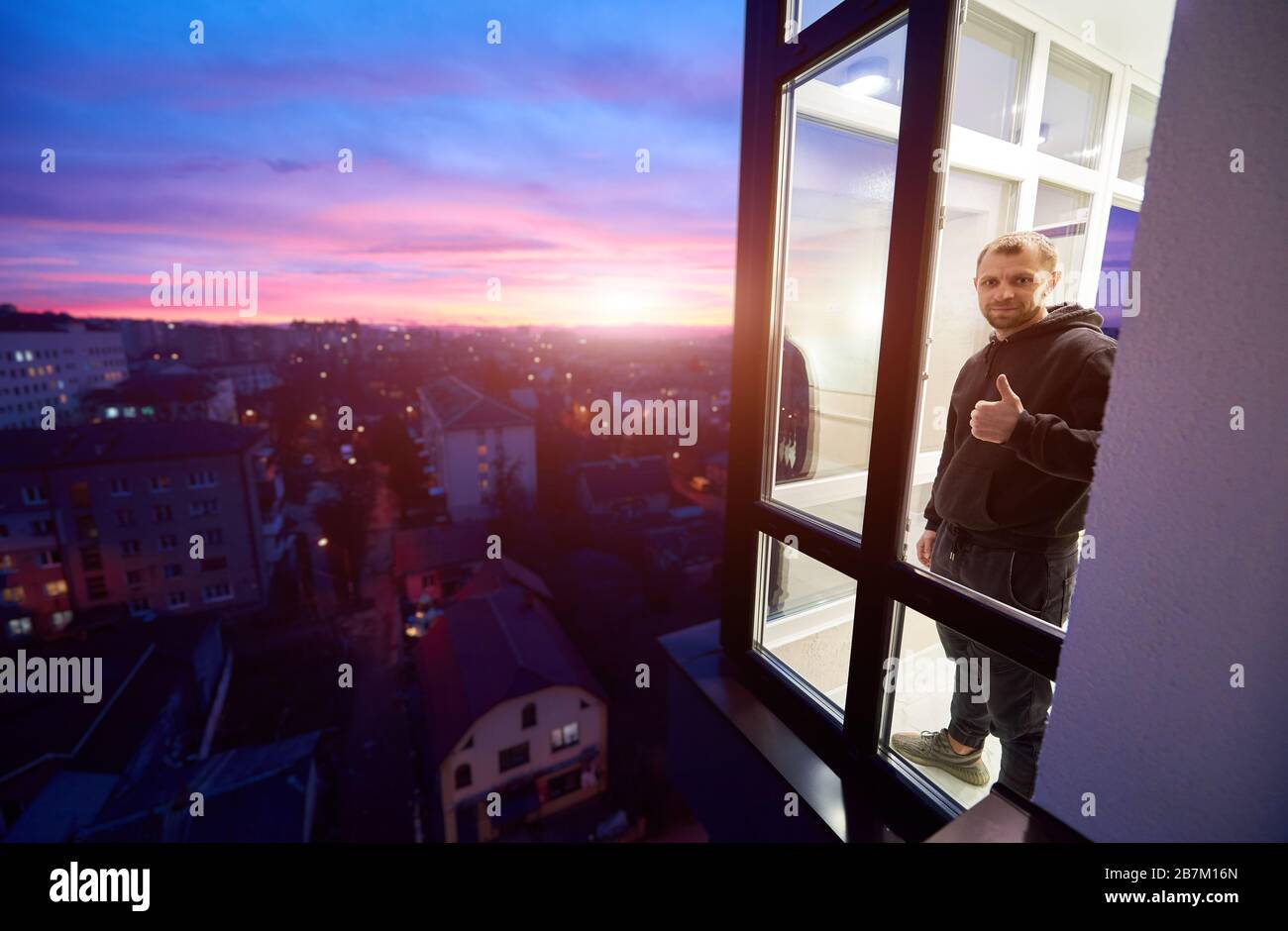 L'homme est debout sur un balcon avec des fenêtres panoramiques et montrant un pouce vers le haut comme s'inscrire avec un coucher de soleil magique à l'extérieur de la fenêtre. Paysage urbain de la hauteur et du ciel multicolore avec soleil couchant Banque D'Images