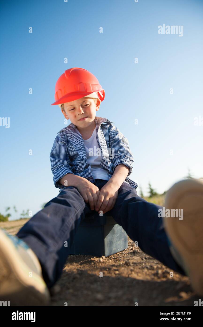 enfant fatigué en casque orange assis sur la boîte à outils après une journée de travail difficile Banque D'Images