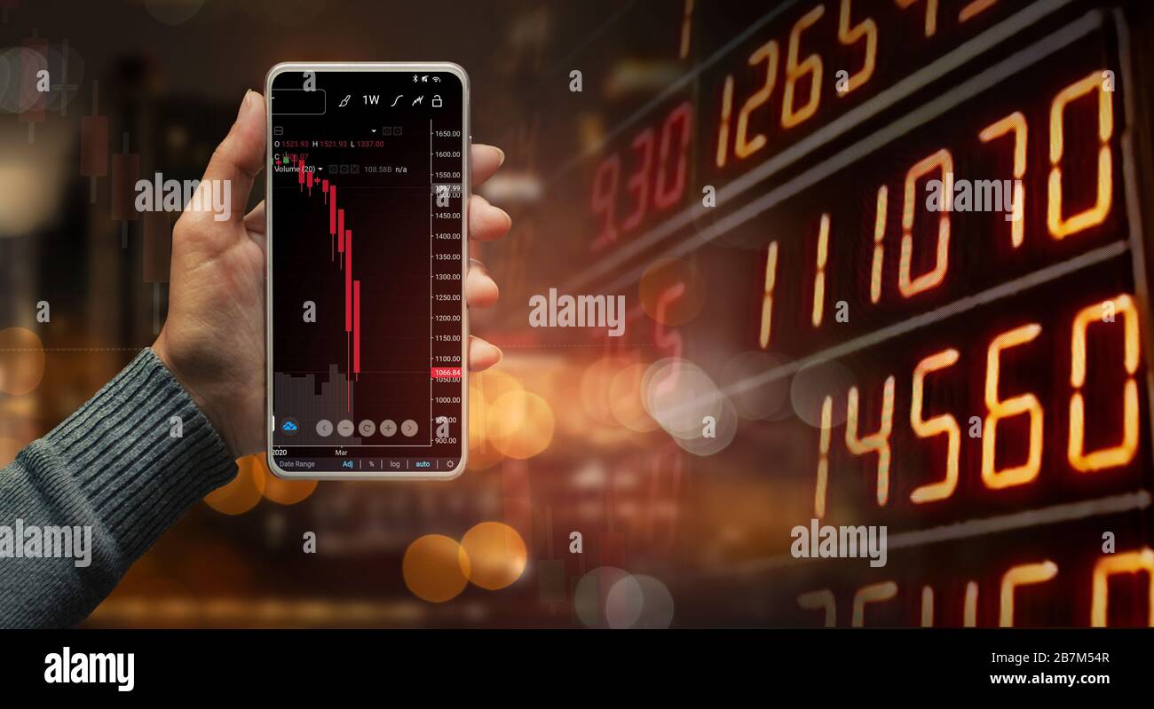 Une main tenant un smartphone noir montrant la tendance des chandeliers du marché boursier sur le panneau d'affichage des données du marché boursier flouté, concept d'affaires et de finances. Banque D'Images
