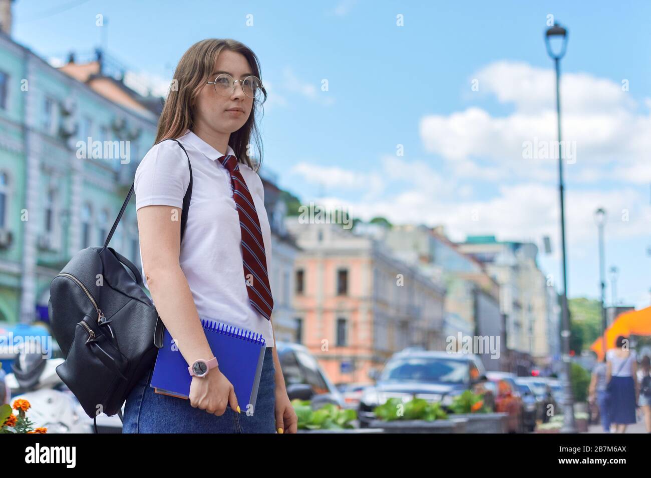 Jeune fille 17, 18 ans étudiant de lycée dans la ville en regardant à côté,  copier l'espace. Femme au niveau de la cravate, lunettes, t-shirt blanc,  sac à dos, ordinateur portable Photo